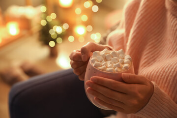 Woman holding cup of hot drink with marshmallows indoors, closeup. Magic Christmas atmosphere