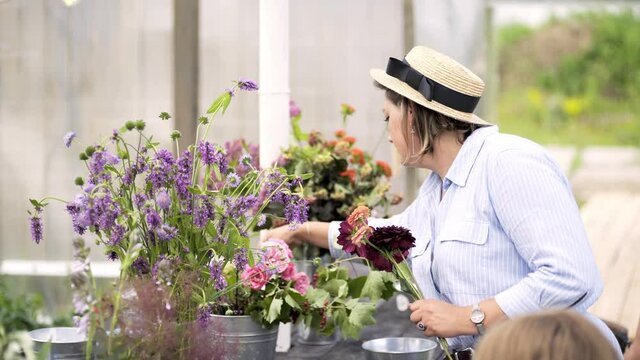 Woman in light blue dress create bouquet with colorful flowers in vases on long wooden table preparing for masterclass in greenhouse or decorate for event celebration