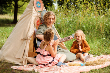 Mom plays the guitar to her children outdoors in summer. Wigwam decoration.