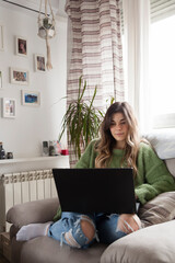 Naklejka premium Lifestyle vertical shot of curly blonde hair styled woman sitting confortable on a sofa while works remote from home using his laptop with home decorated background and heating.