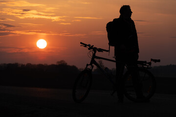Fototapeta premium The silhouette of a cyclist against the background of the sun and the beautiful sky. The outline of a man standing next to his bicycle against the background of the sunset.