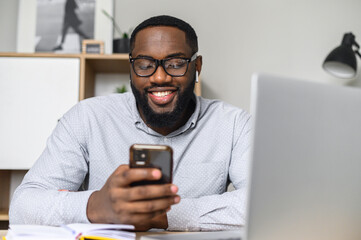 Obraz na płótnie Canvas Happy and positive young African-American adult in glasses sitting at the desk, surfing the Internet on his mobile phone, working on social media projects, checking emails, and chatting with friends