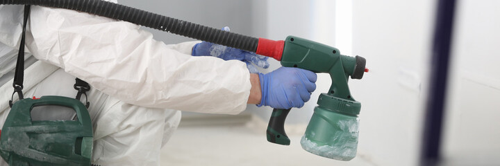 Close-up of professional worker painting wall in grey colour with special spray gun. Person in white protective costume and face mask. Ladder in right side. Renovation and construction site concept