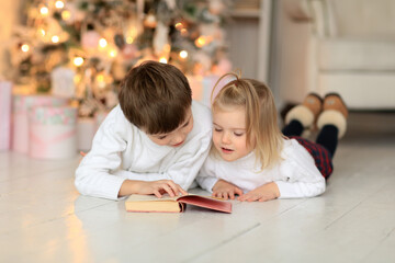 brother and sister in a white sweater on the floor being treated near the Christmas tree and reading a book fun