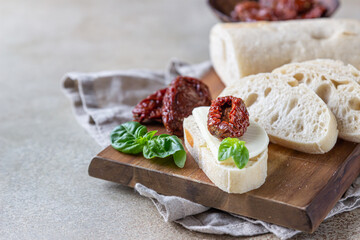 Sliced ciabatta bread with sun-dried tomatoes, cheese, basil and olive oil on wooden board, stone background.