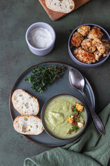 Broccoli cream soup decorated with thyme, cream, roasted cauliflower and fresh bread on green concrete background. Healthy vegetarian food concept. Top view.
