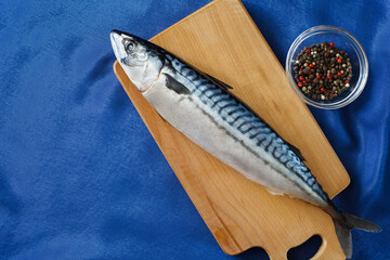 Raw mackerel fish whole, on a cutting board, bowl with a mixture of peppers and spices and a blue satin tablecloth. Top view with copy space.