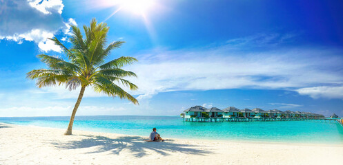 Fototapeta na wymiar Beautiful beach with white sand, turquoise ocean, palm and blue sky with clouds on sunny day. Man sits on sand in shade of palm tree. Sun is at its zenith. Summer tropical landscape, panoramic view.
