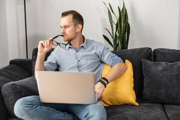 A smart young man took glasses off, sits on the comfortable couch with a laptop and looks away lost...