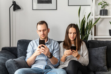 Couple sits on the comfortable couch and using mobile phones separately, a man and a woman lost in...