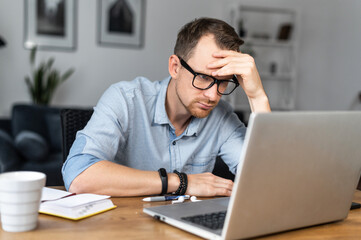 Thoughtful young man in modern glasses and smart casual wear is looking on laptop screen and...