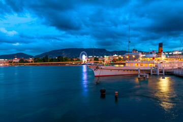 Naklejka premium Dramatic stormy sky of Geneva urban cityscape and Swiss Alps reflecting in Lake Leman, Switzerland in French Swiss. Pier in Quai du Mont-Blanc ferry terminal at dusk, illuminated in the evening.
