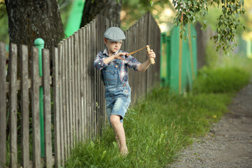 little boy in a cap and a plaid shirt, denim shorts and shooting a slingshot