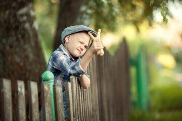 little boy a bully in a cap and a plaid shirt sitting on a wooden fence shooting a slingshot