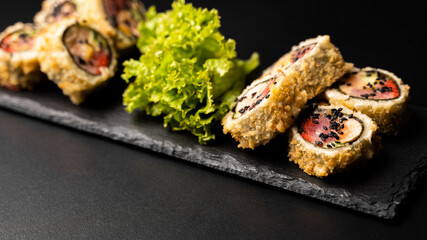Custom sushi roll in tempura with nori, fresh salmon, tuna, avocado, masago caviar, drizzled with pineapple sauce with salad pouring as decoration on a black plate on a black table and background.