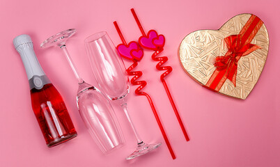 Two champagne glasses with red plastic drinking straws with hearts, gift box and red champagne bottle on a pink background.