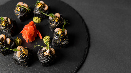 Fototapeta premium Sushi roll with nori, black rice, crab meat, cucumber, avocado, smoked salmon mousse, oar caviar, masago, shrimp cocktail and edible gold leaf with red ginger and wasabi on black table and background.