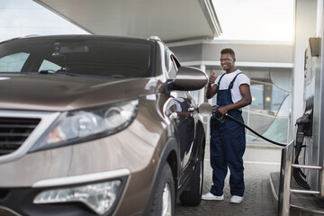 Gas station, transportation and energy concept. African man, professional gas station worker in uniform, smiling and showing thumb up to camera, while refueling modern car