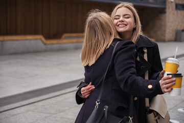 Fototapeta na wymiar Two blonde adult caucasian women meet in cold dark evening. They wear coats and holds to go cup of coffee in their hands. They laugh and smiles happily together. Space for text