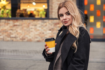 Blonde woman in dark coat drinks coffee in to go cup. Dark cold autumn evening. Europeaan city lights on the background. Space for text