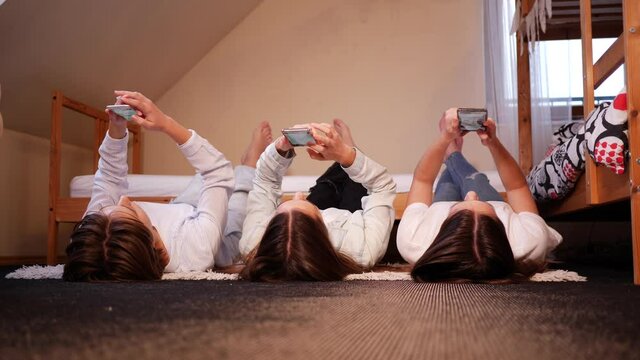 Three triplet sisters teenager girls at home use smartphones lie on floor in bedroom spend time in social media