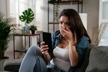 Fototapeta premium Sad woman sitting on the couch, using the phone. Upset woman waiting for a phone call.