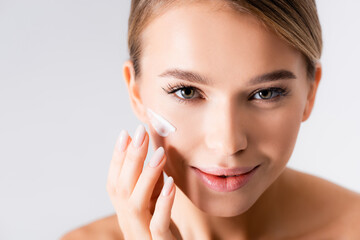 joyful young woman applying moisturizing cream on face isolated on white