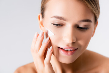 young woman with bare shoulders applying moisturizing cream on face isolated on white