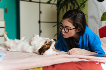Happy girl in eyeglasses stroking her lovely dog while lying on bed