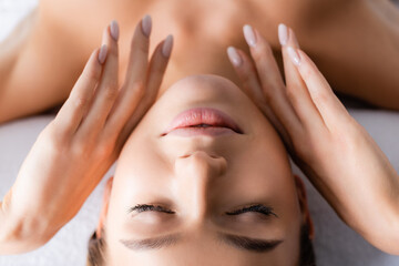 Young woman with closed eyes touching face and lying on massage table in spa salon