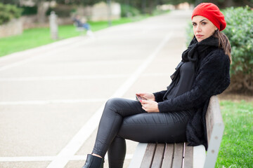 Fototapeta premium Ragazza mora vestita di nero con cuffietta rossa è seduta in u panchina in un parco