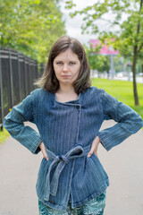 An upset girl stands with her hands on her hips on the street against the background of the park...