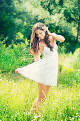 Naklejka na ściany i meble Smiling young woman in white dress with flower against background of summer green park. Beautiful healthy happy girl enjoying freedom outside in nature.