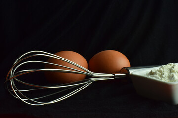 Whisk rod, kitchen utensil on black background, eggs and flour.