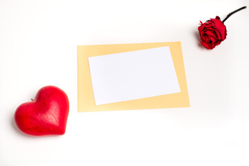Top view on a white and blank gift card on a sand-colored envelope, with a red love heart on its left and a red rose on the right, on a white background. Valentine's day and love 