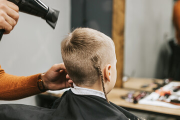 Fototapeta premium the process of hair styling a blond boy with with hair dryer and comb in a chair in a barbershop salon, a barbershop concept for men and boys