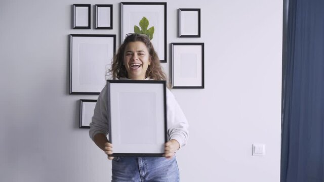 Excited young brunette with long curly hair dances jumping and hiding behind wooden picture frame against decorated wall under bright light in new flat