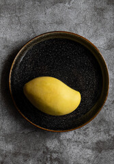 One yellow mango in a dark plate on a concrete background in vertical orientation. Copy space, top view. Tropical fruit 