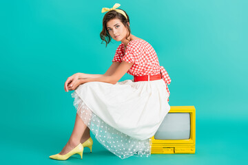 pretty woman in retro clothes looking at camera while sitting on yellow vintage tv set on turquoise