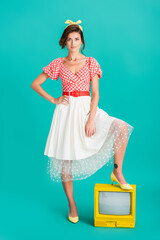stylish pin up woman with hand on hip looking at camera while stepping on yellow vintage tv on...