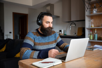 Young man at home at table working on computer with headphones - Concept of smartworking in lockdown - Millennials in a business meeting with his colleagues remotely - Male attends an online course