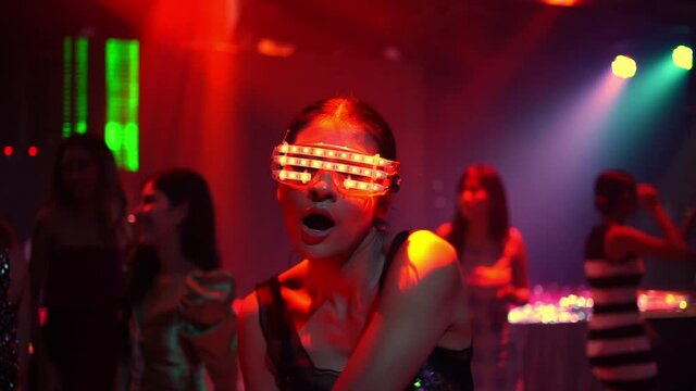 Confidence Asian woman wear LED glowing glasses dancing to the music played by dj with multi-color illuminated neon night lights at night club. Group female enjoy nightlife and having fun dance party.