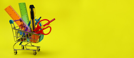 Shopping cart with stationery, school supplies on yellow background