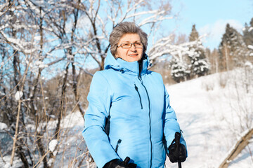 Elderly woman in glasses with Nordic walking poles on the background of winter forest. Hike retirement concept. Sunny winter day.