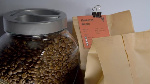 Closeup POV shot of a man's hands using sprung metal clips to attach descriptive labels from the roaster to fresh, sealed packs of an espresso blend of coffee beans, next to a full jar.