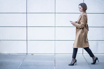 bella manager con camicia bianca e cappotto in contesto urbano , cammina guardando il suo smartphone