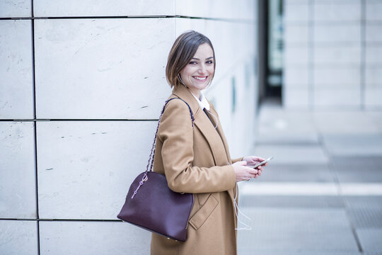 bella manager con camicia bianca e cappotto in contesto urbano , sorride con in mano il proprio smartphone