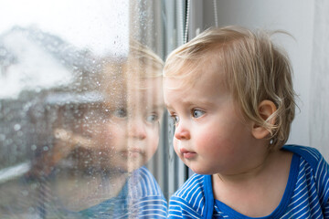 Sad little child sitting in front of window and looking out in rainy day. Stay at home coronavirus...