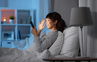 Fototapeta na wymiar technology, internet, communication and people concept - happy smiling young asian woman with smartphone having video call lying in bed at home at night