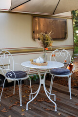 Chairs and table with tea set placed outside cozy retro caravan trailer on lawn. Interior cozy wooden RV house porch with garden furniture. Decor outdoor yard. Caravan trailer parked in camping site. 